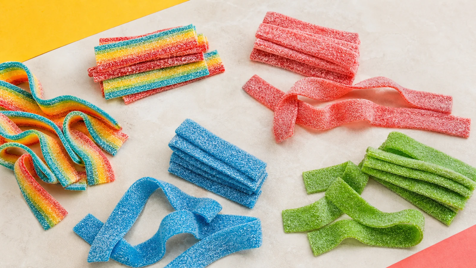 Rainbow, berry, blue raspberry, and green apple sour gummy belts on a cream surface.